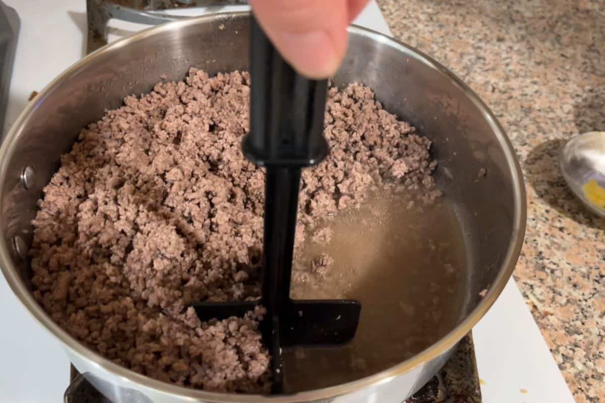 A hand uses a black meat chopper to break up cooked ground beef simmering in a pot on a stovetop, with liquid visible at the bottom and a granite countertop nearby.