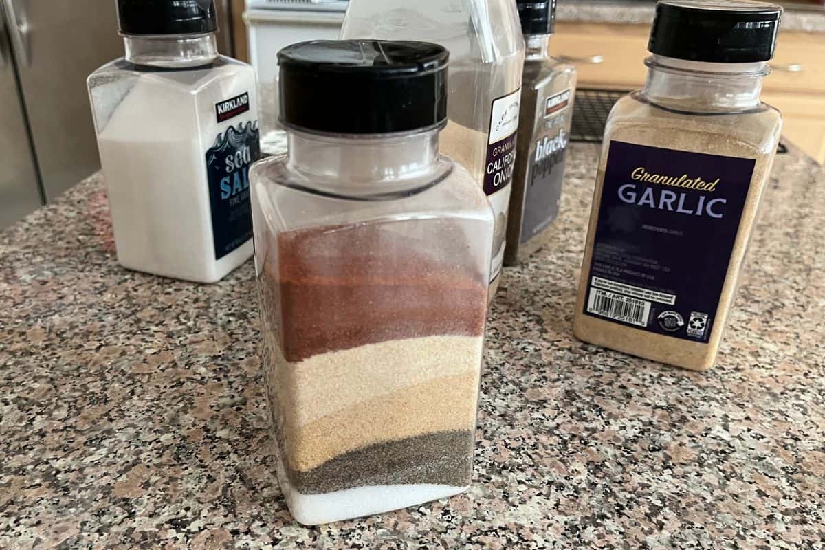 A clear spice container filled with neatly layered seasonings sits on a granite countertop, surrounded by other labeled spice bottles including sea salt, granulated garlic, and onion powder.