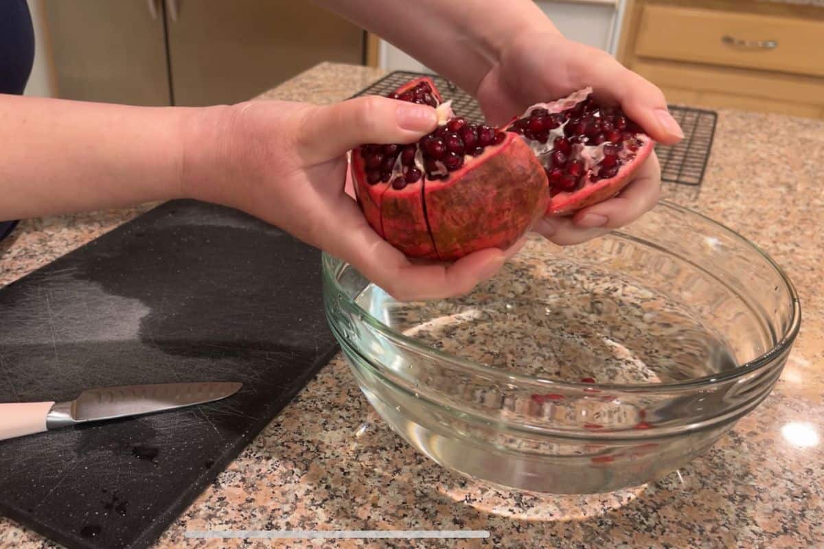 A person breaks open a pomegranate over a glass bowl of water on a granite countertop. A knife rests on a black cutting board nearby.