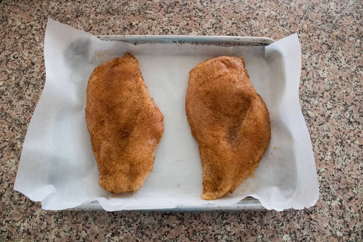 Two raw chicken breasts coated with seasoning are placed on a sheet of parchment paper in a baking tray, set on a speckled countertop.