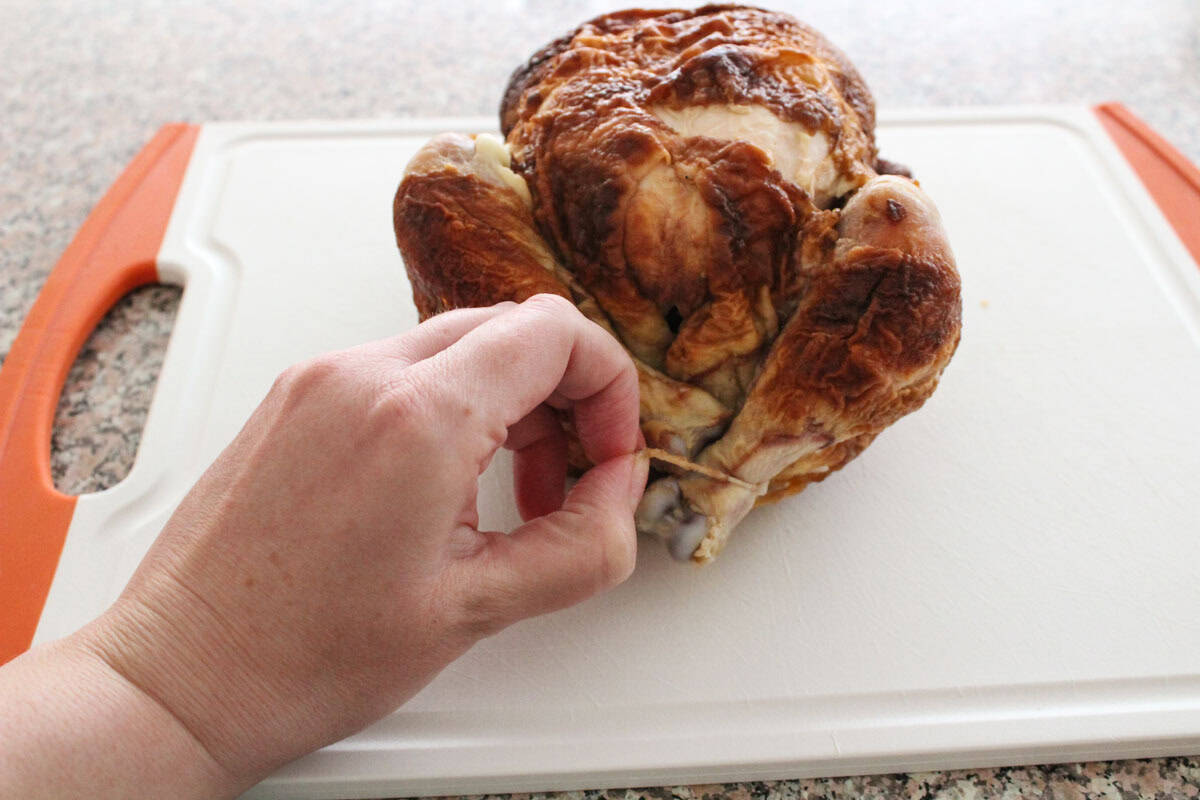 A hand unties kitchen twine from the legs of a cooked rotisserie chicken resting on a white cutting board with orange handles.