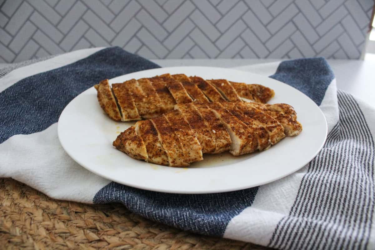 Two sliced, seasoned chicken breasts on a white plate, placed on a blue and white striped towel with a woven placemat underneath and a herringbone-patterned background.