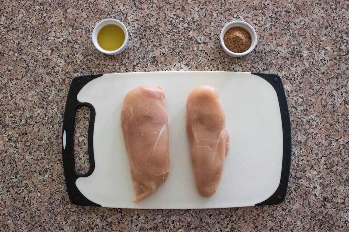 Two raw oven baked chicken breasts on a white cutting board with a black handle, set on a speckled countertop. Above them sit two small bowls—one filled with olive oil and the other with a brown spice mix.