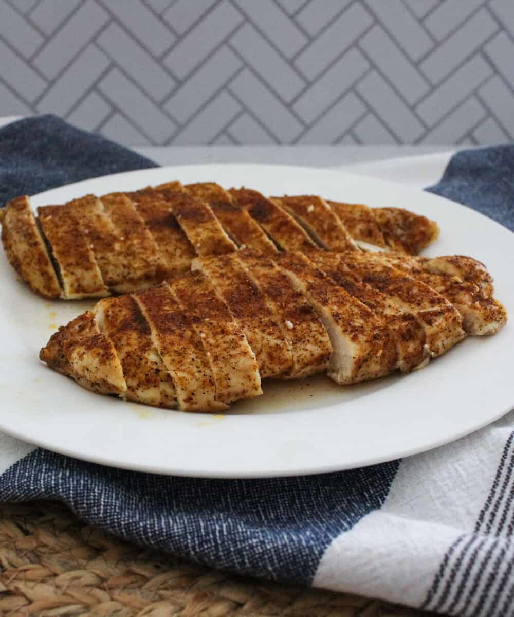 Two sliced, oven baked chicken breasts are arranged on a white plate placed on a striped kitchen towel, with a woven mat and a gray patterned wall in the background.