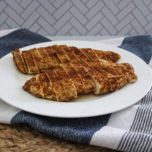 Two sliced, oven baked chicken breasts are arranged on a white plate placed on a striped kitchen towel, with a woven mat and a gray patterned wall in the background.