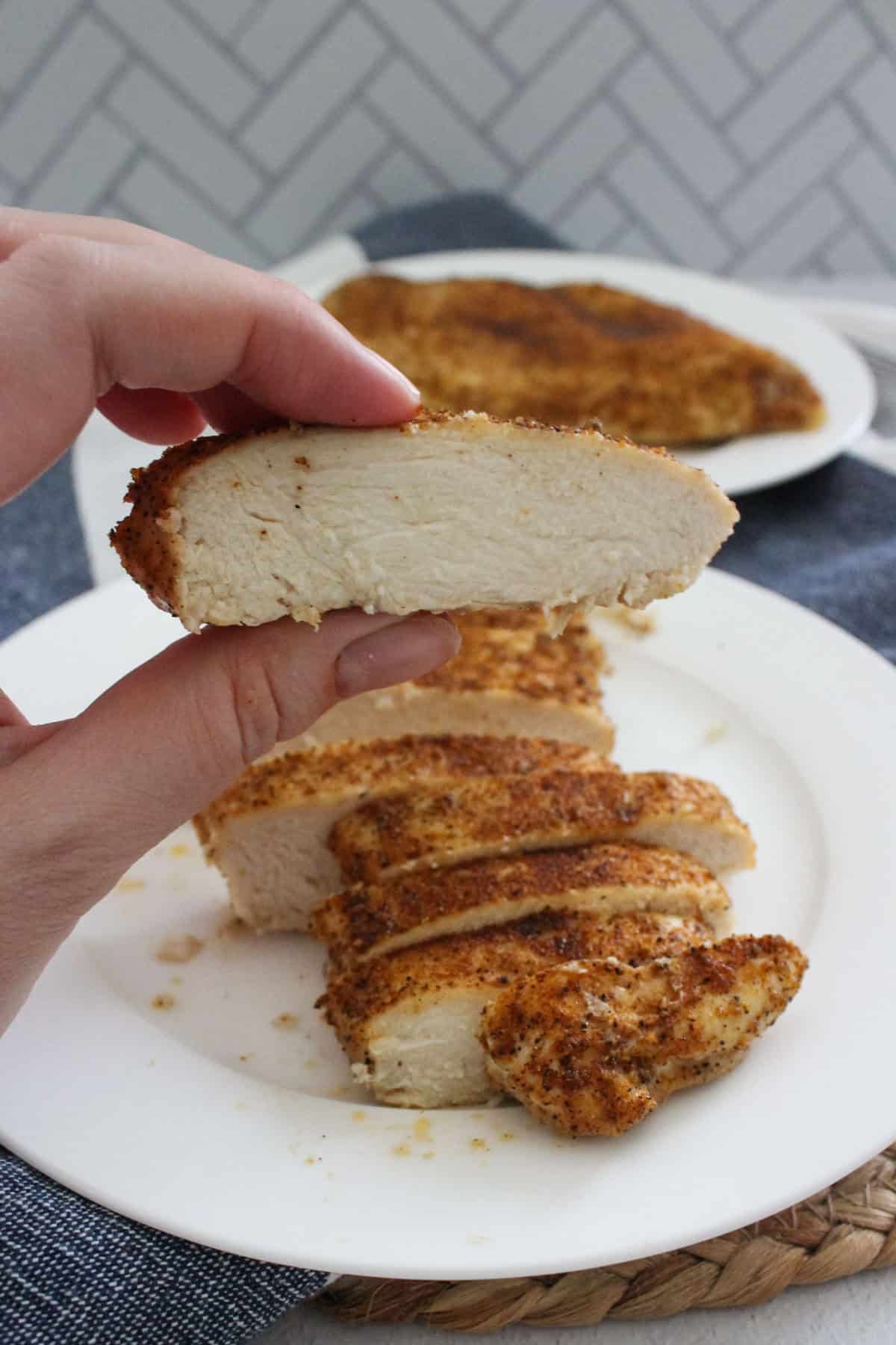 A hand holds up a slice of seasoned, cooked chicken breast to show its juicy interior, with more sliced chicken arranged on a white plate in the background.