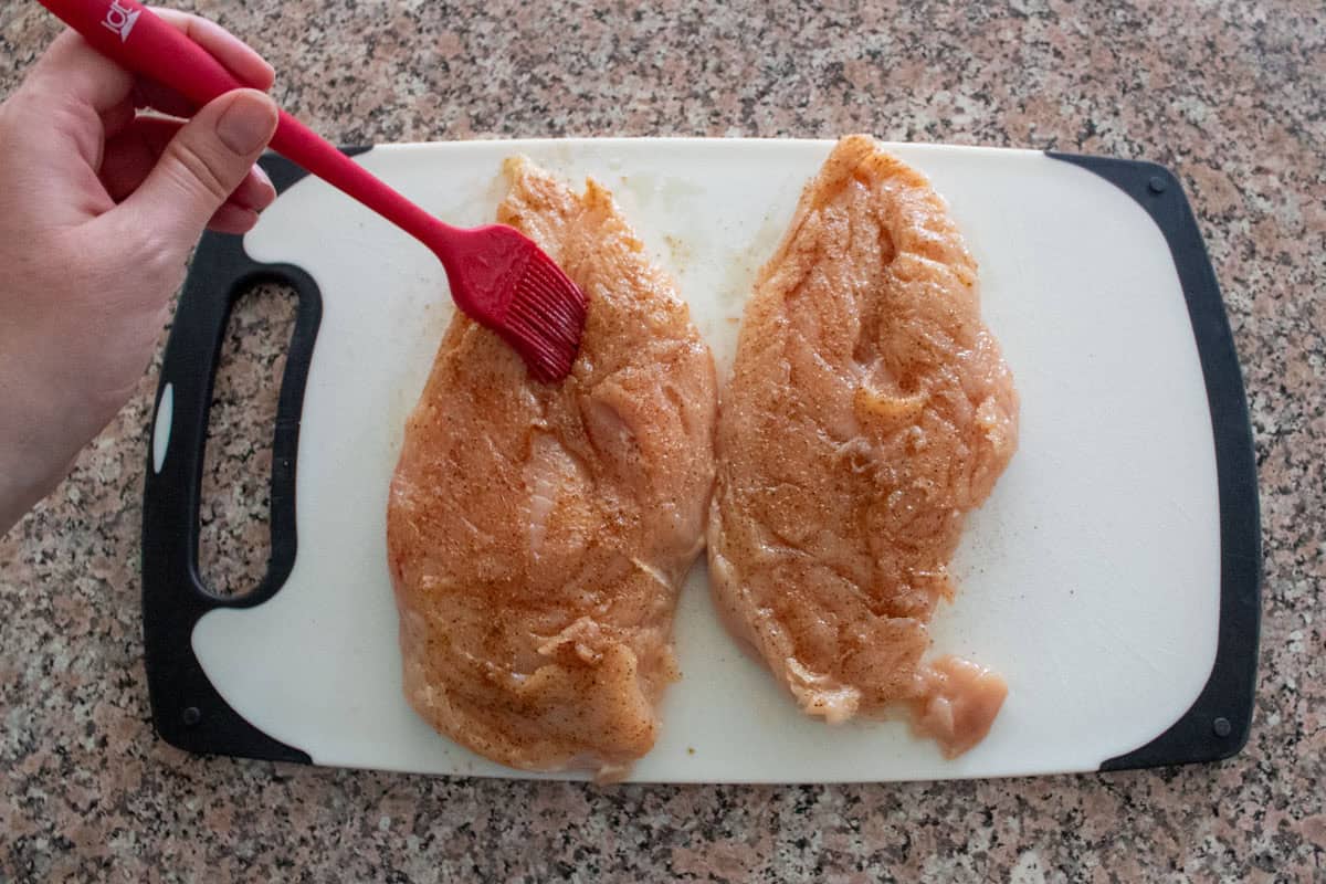 A hand uses a red brush to spread seasoning on two raw, oven baked chicken breasts placed on a white cutting board with a black handle on a speckled countertop.