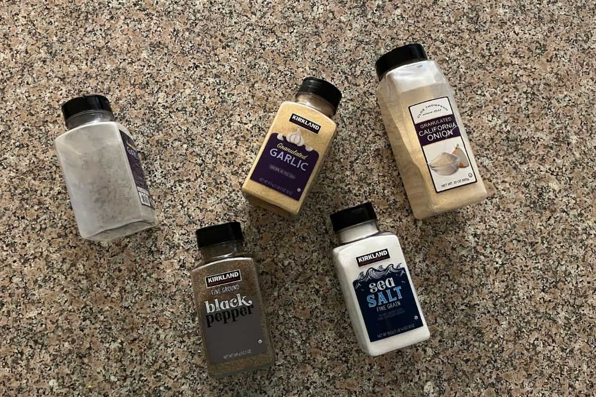 Five Kirkland spice containers (sea salt, black pepper, garlic powder, onion powder, and an empty container) are arranged on a brown speckled countertop.