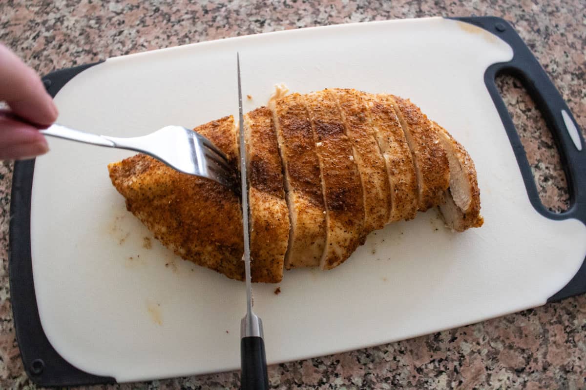 A hand uses a fork and knife to slice a seasoned, oven baked chicken breast on a white cutting board with black handles, set on a speckled countertop.