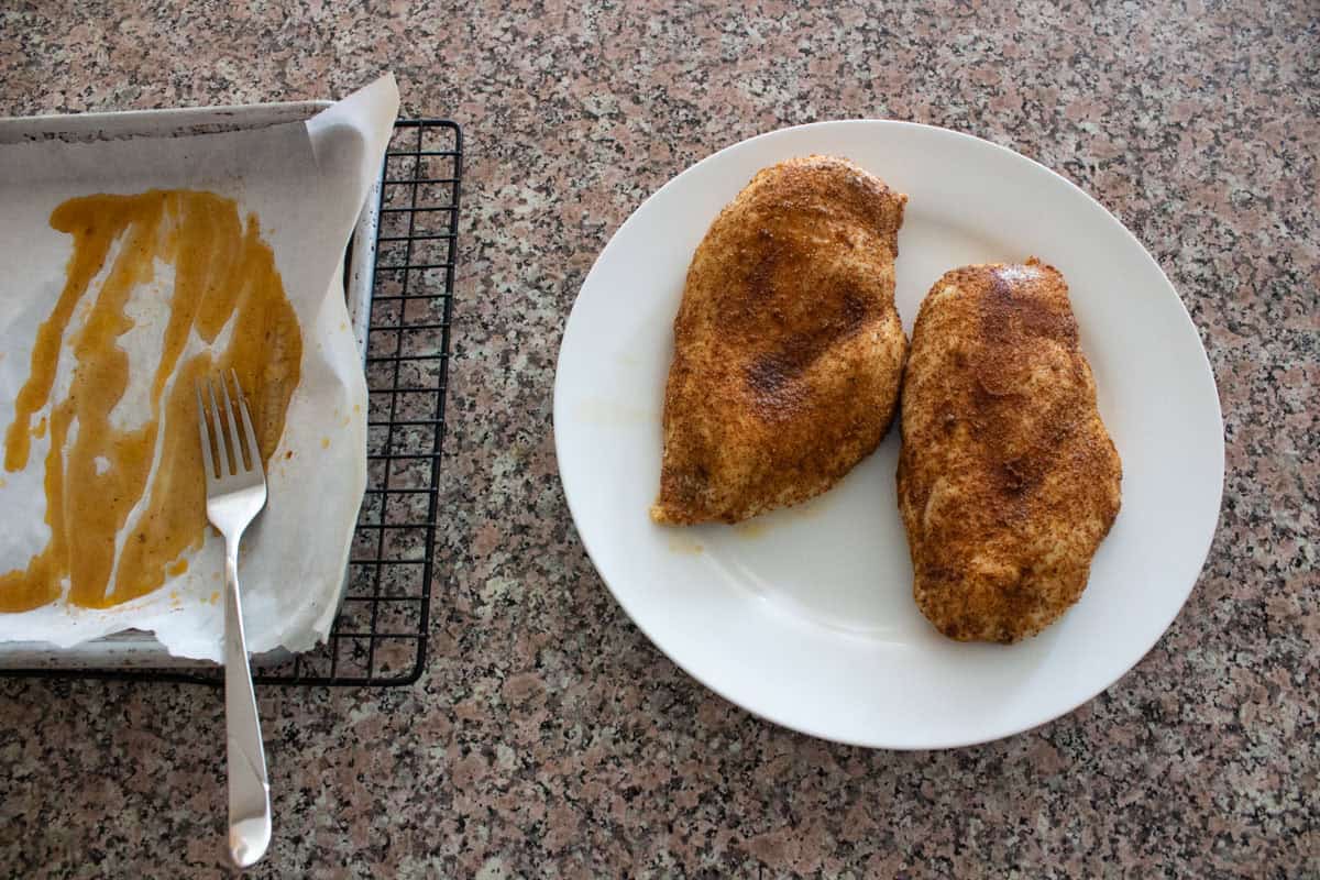 Two seasoned, cooked chicken breasts on a white plate next to a baking tray lined with parchment paper, showing leftover seasonings and a fork, all on a speckled countertop.