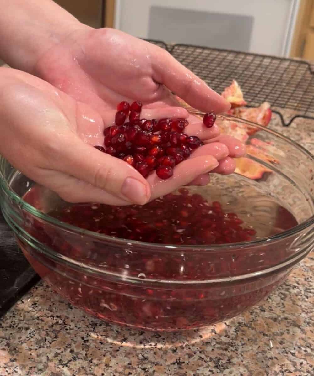 Two hands hold freshly deseeded pomegranate arils over a glass bowl filled with water and more arils on a granite countertop. Pieces of pomegranate rind are visible in the background.