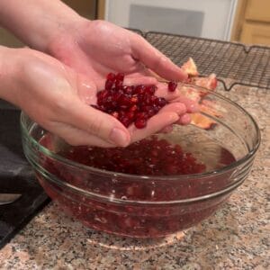 Two hands hold freshly deseeded pomegranate arils over a glass bowl filled with water and more arils on a granite countertop. Pieces of pomegranate rind are visible in the background.