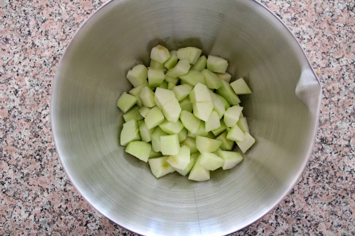 A stainless steel mixing bowl containing peeled and chopped green apple pieces, placed on a speckled countertop.