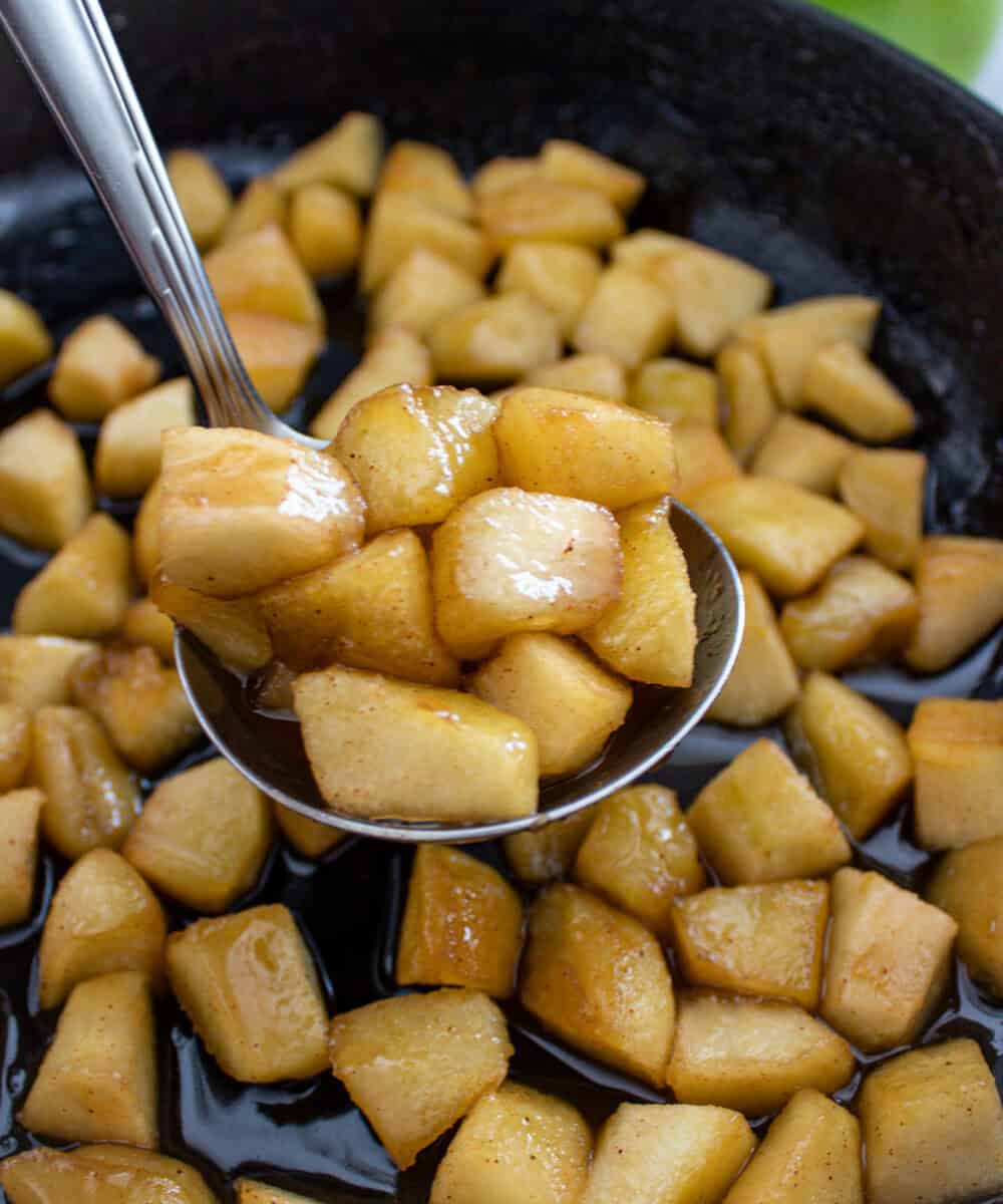 A close-up of a spoon lifting caramelized diced apples in syrup from a skillet, with more apple pieces and caramel sauce visible in the pan.