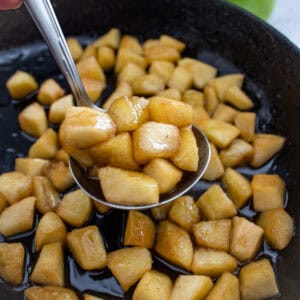 A close-up of a spoon lifting caramelized diced apples in syrup from a skillet, with more apple pieces and caramel sauce visible in the pan.