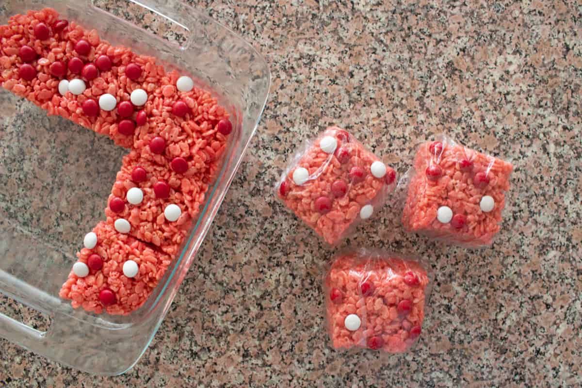 A glass dish with pink rice crispy treats topped with red and white candies, next to three wrapped individual squares of the same treat, all on a granite countertop.
