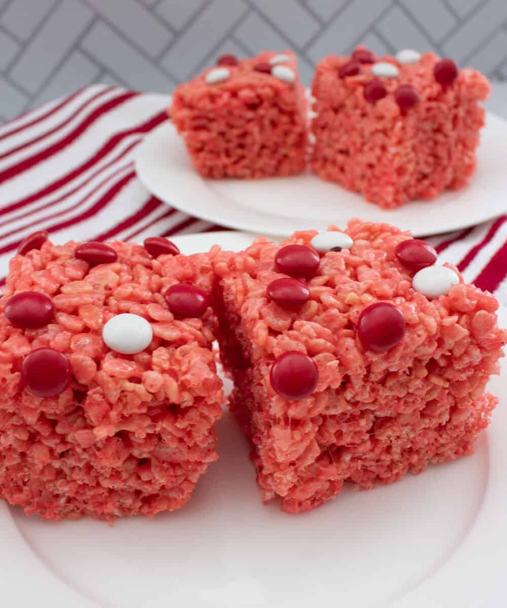 Two red Rice Krispies treats topped with red and white candy dots are on a white plate in the foreground, with more treats on a second plate in the background. A red and white striped towel is nearby.