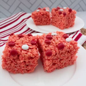Two red Rice Krispies treats topped with red and white candy dots are on a white plate in the foreground, with more treats on a second plate in the background. A red and white striped towel is nearby.