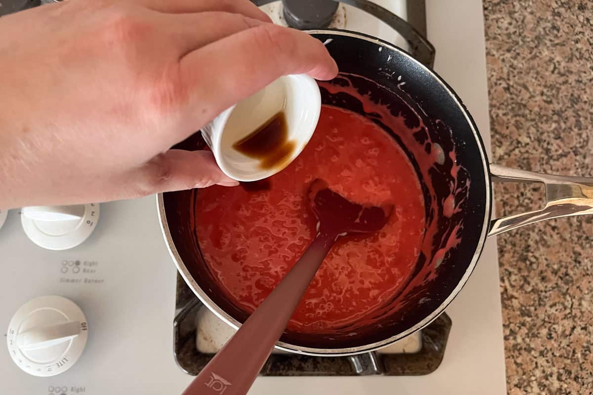 A hand pours a small cup of vanilla extract into a saucepan with a thick red mixture being stirred on a stove.