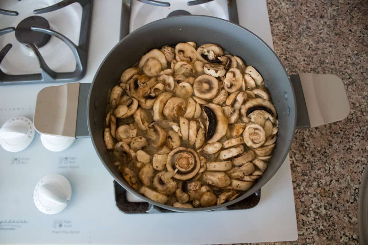 A pot filled with sliced mushrooms cooking on a stovetop burner, with stove knobs and a speckled countertop visible nearby.