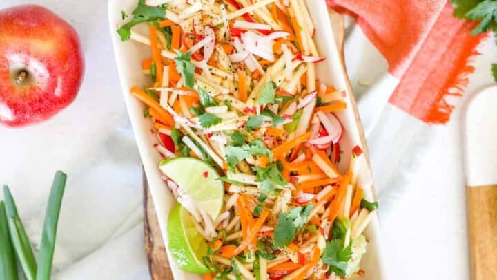 A rectangular white dish filled with a colorful salad of julienned apples, carrots, and radishes, garnished with fresh cilantro and lime wedges. An apple, green onions, and a red napkin are nearby on the white surface.