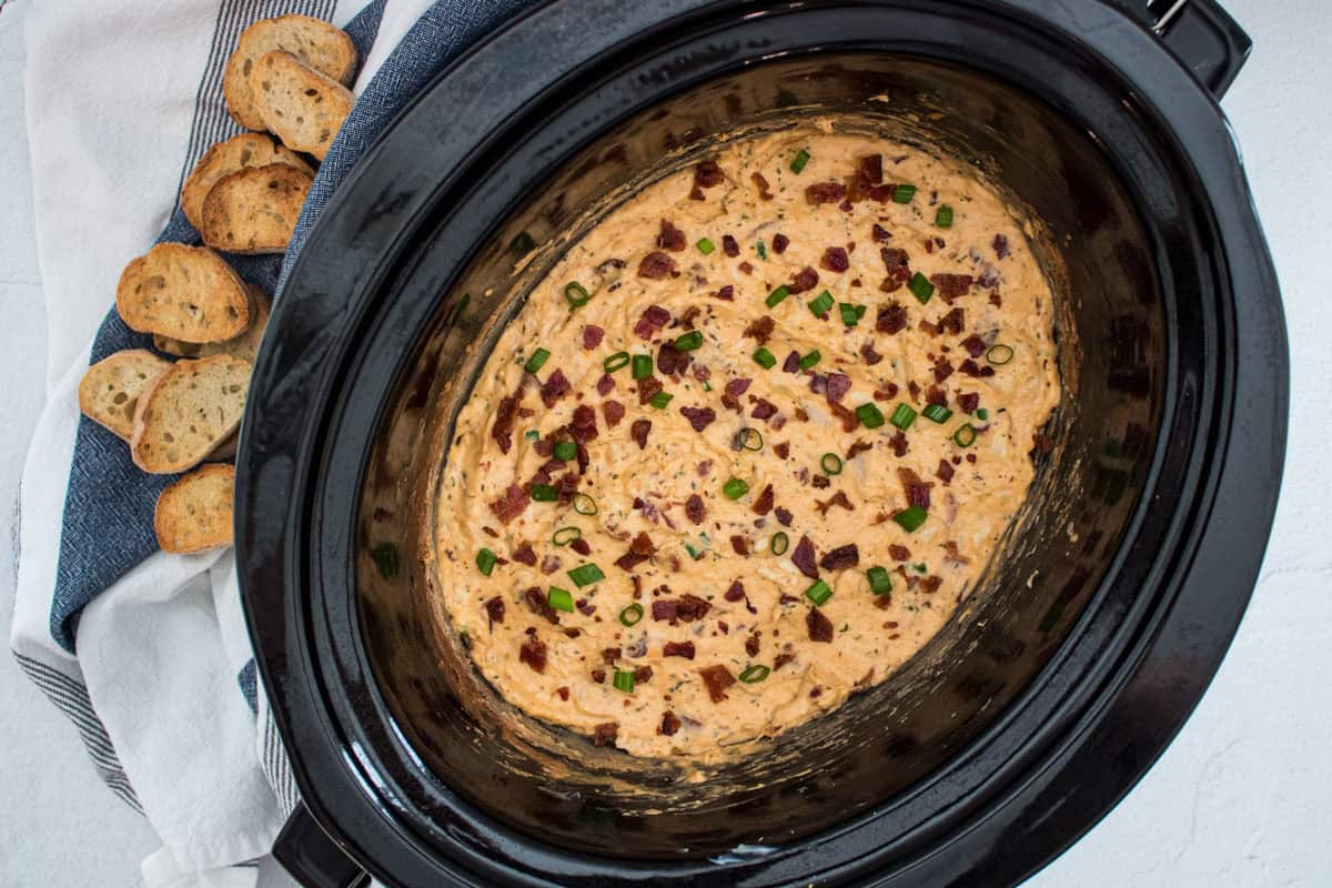 A slow cooker filled with a creamy dip topped with chopped bacon and green onions, next to a white towel holding several slices of toasted baguette.