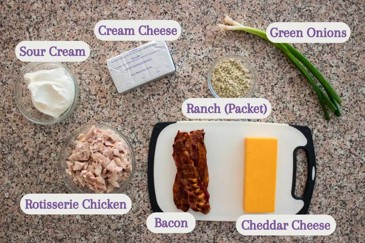 Ingredients for a recipe are laid out on a countertop: sour cream, cream cheese, green onions, ranch packet, rotisserie chicken, bacon, and a block of cheddar cheese, each labeled.
