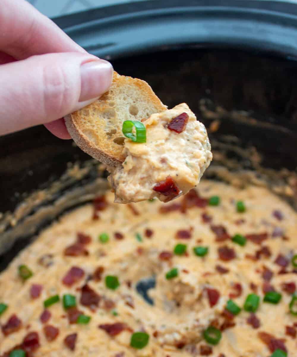 A hand holds a slice of bread dipped in a creamy bacon and cheese dip, garnished with chopped green onions, over a slow cooker filled with more of the dip.