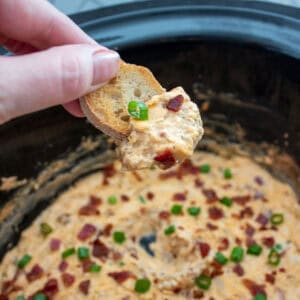 A hand holds a slice of bread dipped in a creamy bacon and cheese dip, garnished with chopped green onions, over a slow cooker filled with more of the dip.