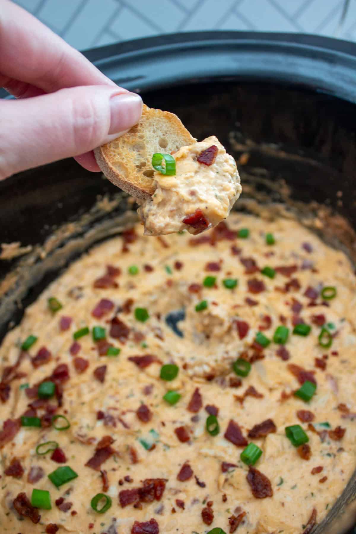 A hand holds a small toasted bread slice topped with creamy dip, garnished with chopped green onions and bacon bits, over a slow cooker filled with the same dip.