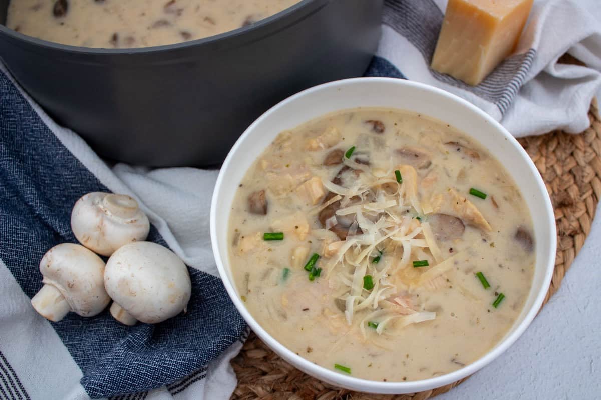 A bowl of creamy mushroom soup topped with grated cheese and chopped chives sits on a woven mat, next to fresh mushrooms, a pot of soup, and a block of cheese.
