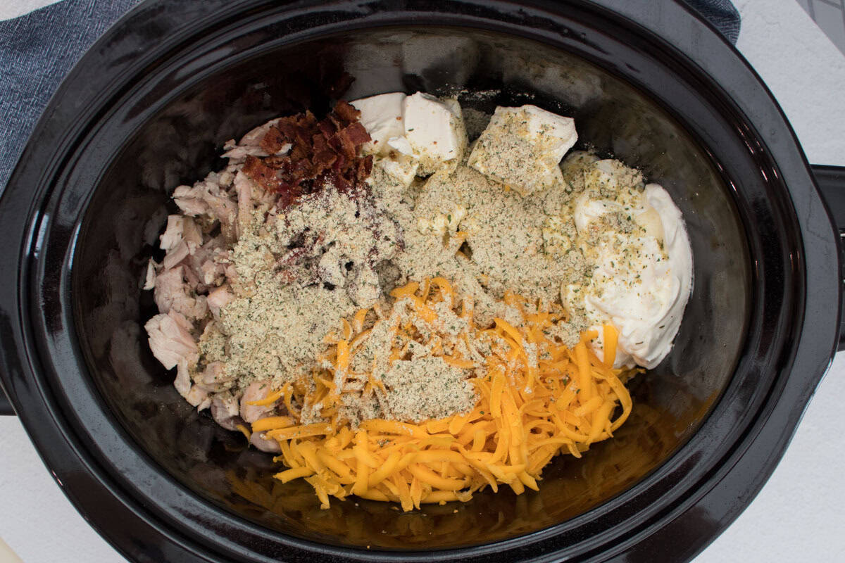 A slow cooker filled with shredded chicken, crispy bacon bits, shredded cheddar cheese, cream cheese, and herb seasoning before being mixed together.