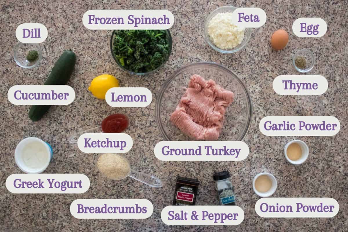 A top view of various labeled ingredients on a countertop, including ground turkey in a bowl, frozen spinach, feta, egg, cucumber, lemon, dill, thyme, ketchup, garlic powder, Greek yogurt, breadcrumbs, salt, pepper, and onion powder.