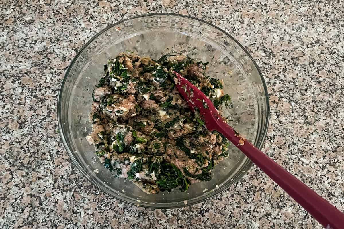 A glass bowl filled with a mixture of cooked greens, browned meat, and white cheese on a speckled countertop. A red mixing spoon rests in the bowl.