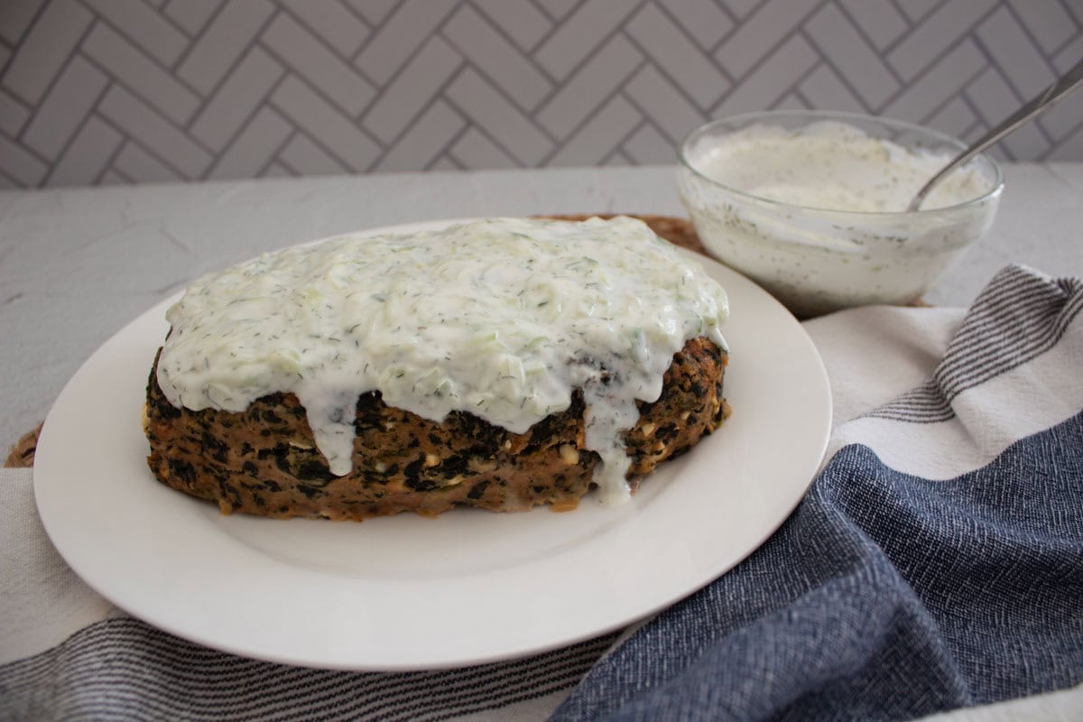 A turkey meatloaf with spinach topped with creamy white sauce on a white plate, with a bowl of extra sauce and a spoon beside it, set on a light table with a blue-striped napkin.