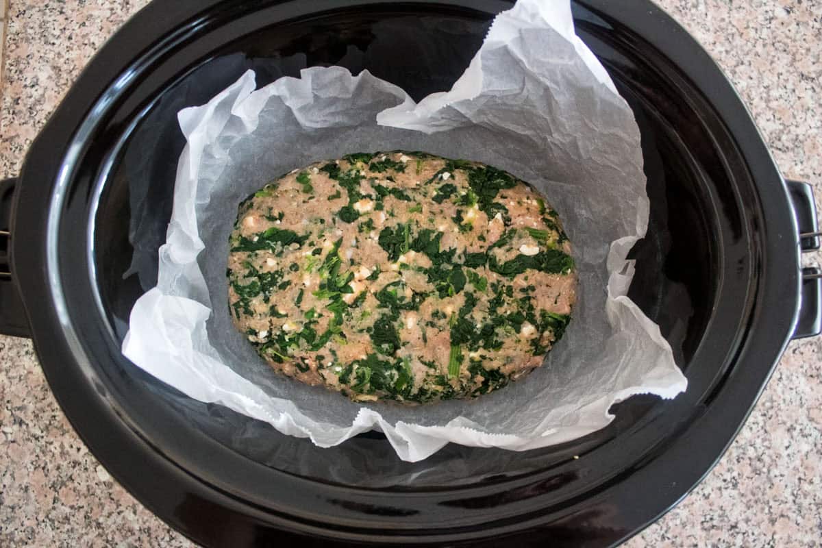A raw spinach and meat mixture is shaped into a loaf and placed on parchment paper inside a black slow cooker, ready to be cooked. The slow cooker sits on a speckled countertop.
