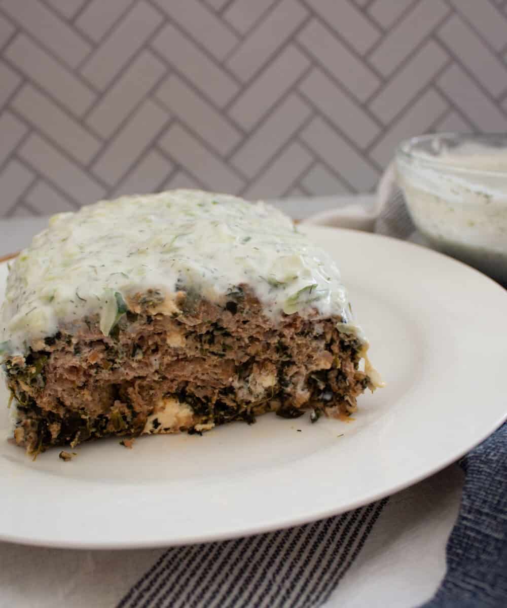 A rectangular slice of meatloaf covered in creamy white sauce sits on a white plate. A bowl of extra sauce and a striped cloth are nearby, with a herringbone-patterned background.