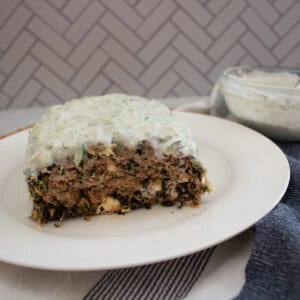 A rectangular slice of meatloaf covered in creamy white sauce sits on a white plate. A bowl of extra sauce and a striped cloth are nearby, with a herringbone-patterned background.
