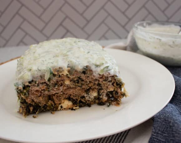 A rectangular slice of meatloaf covered in creamy white sauce sits on a white plate. A bowl of extra sauce and a striped cloth are nearby, with a herringbone-patterned background.