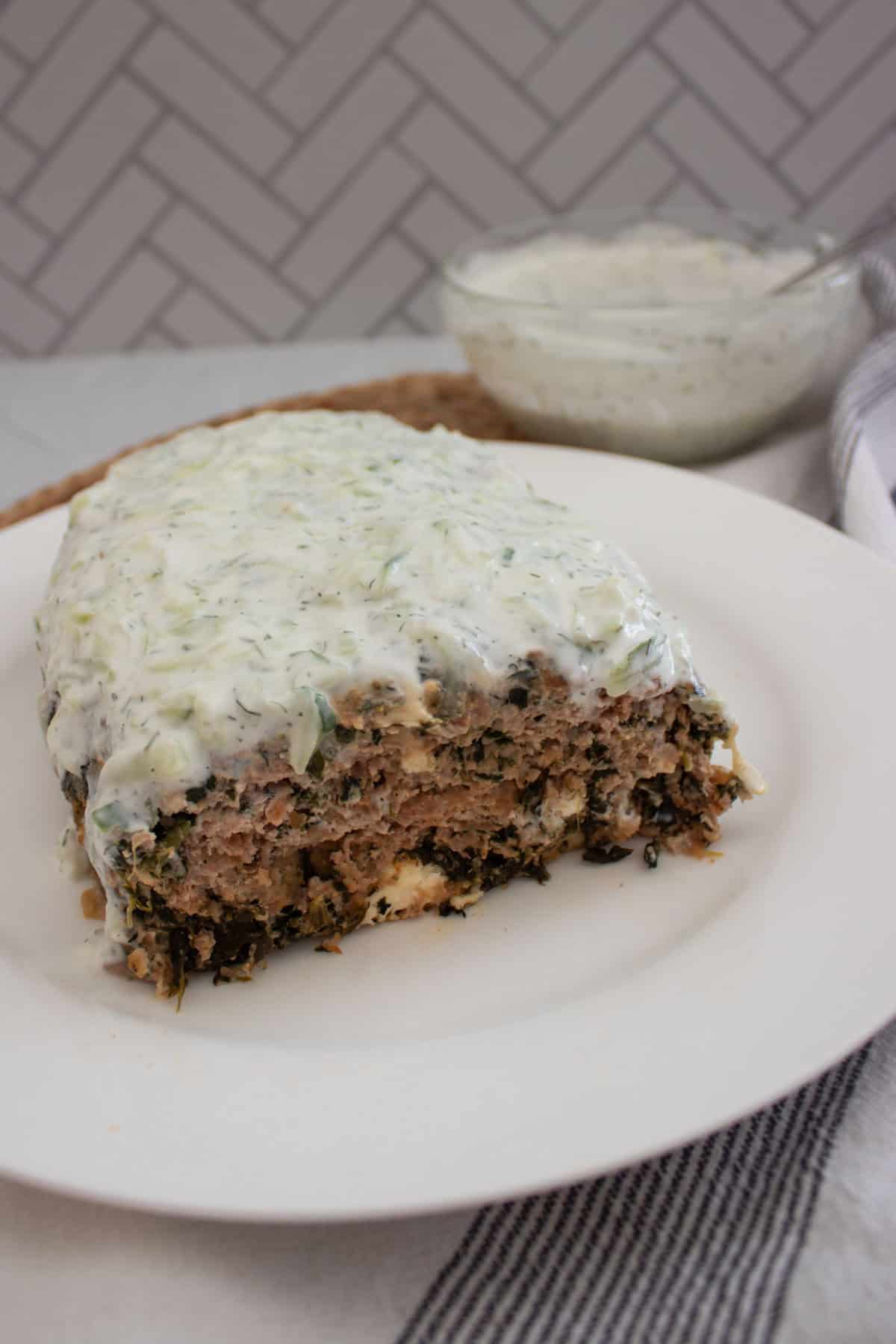 A sliced meatloaf filled with herbs and greens, topped with a creamy white sauce, is served on a white plate. A bowl of extra sauce and a striped napkin are in the background.