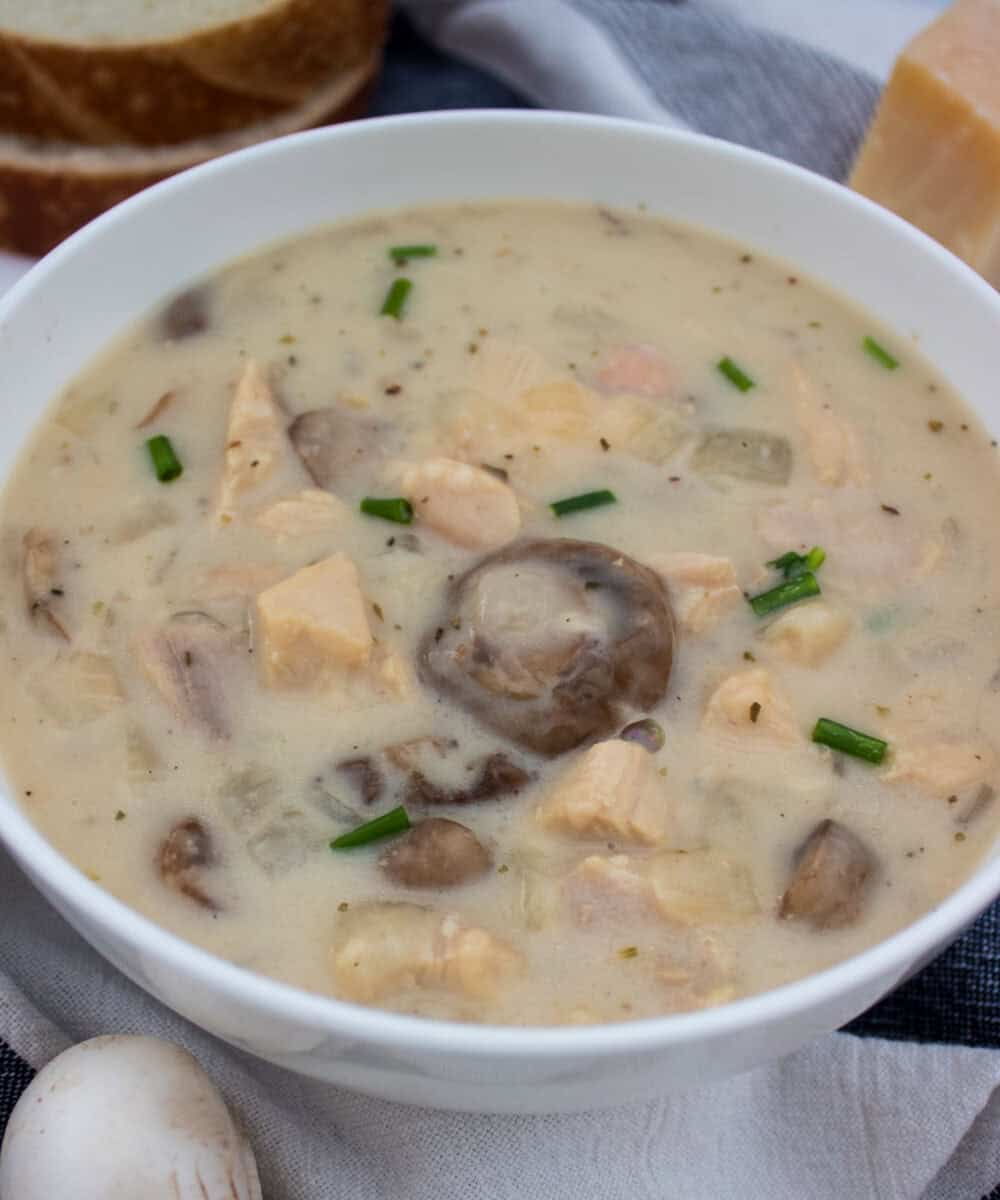 A bowl of creamy soup with chunks of chicken, sliced mushrooms, and chopped chives, placed on a napkin with bread slices and a block of cheese in the background.