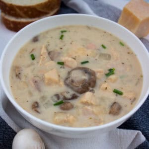 A bowl of creamy soup with chunks of chicken, sliced mushrooms, and chopped chives, placed on a napkin with bread slices and a block of cheese in the background.