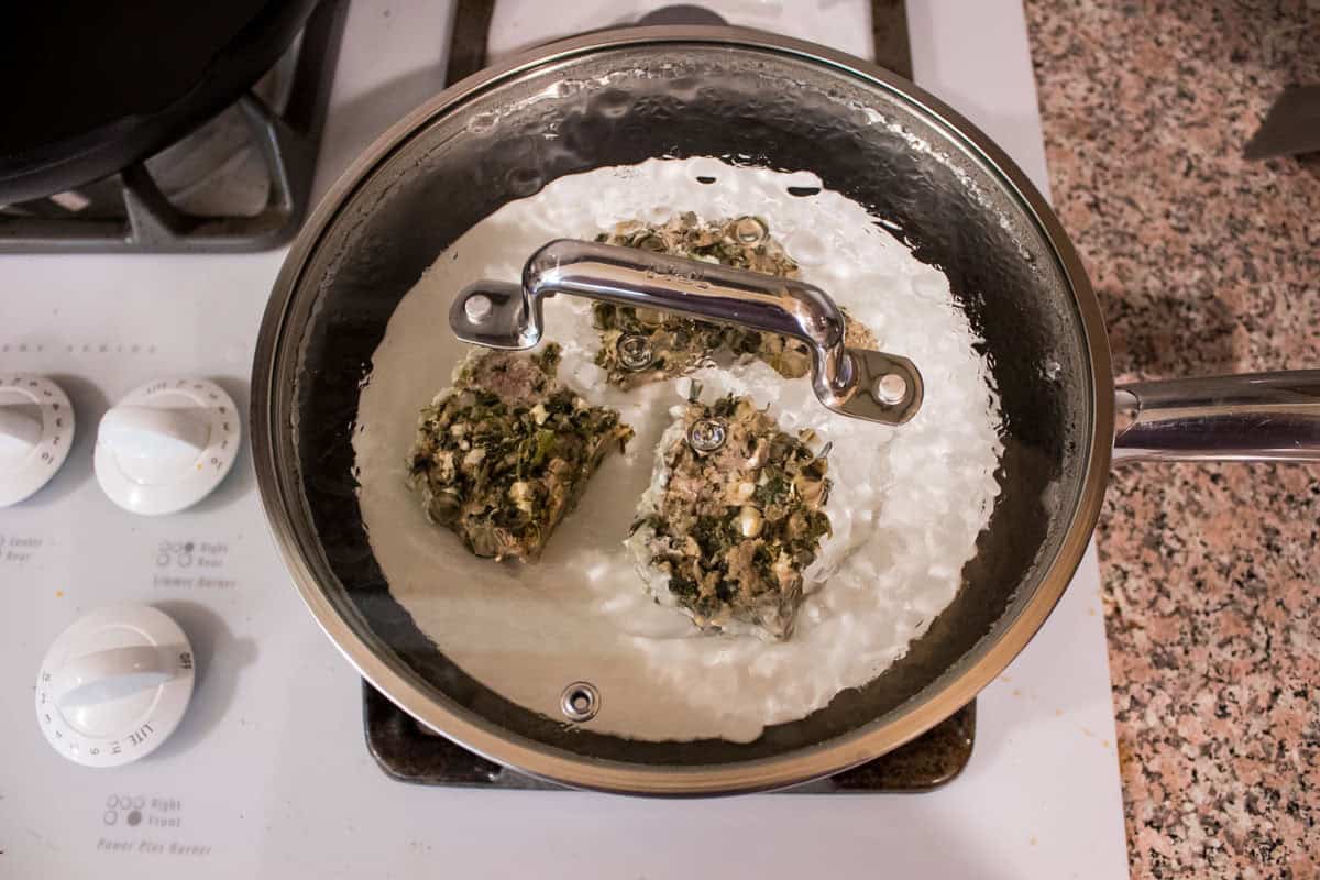 A covered pan on a stove contains three pieces of turkey meatloaf on a heat safe plate. The pan’s transparency reveals the contents, and stove knobs and a speckled countertop are visible nearby.