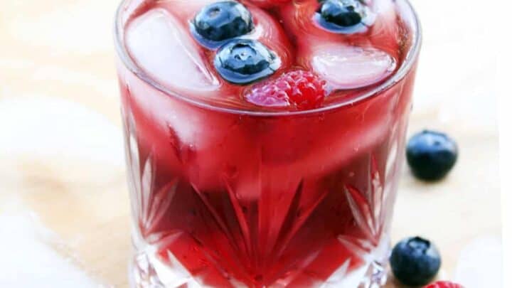 A glass of red iced beverage garnished with blueberries and raspberries, surrounded by ice cubes and more berries on a light wooden surface.