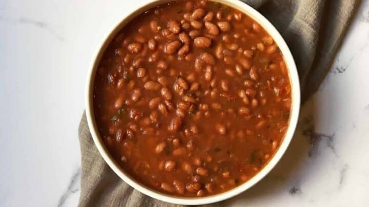 A white bowl filled with baked beans in sauce sits on a light gray cloth, placed on a white marble surface.