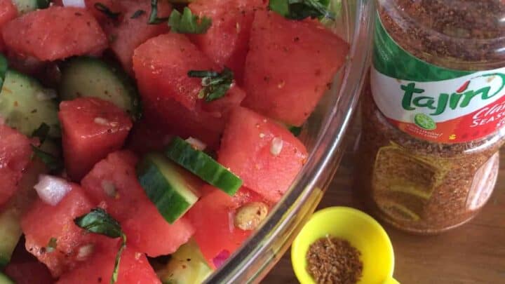 A bowl of watermelon and cucumber salad garnished with herbs sits next to a bottle of Tajín seasoning, a halved lime, and a yellow measuring spoon with seasoning on a wooden surface.