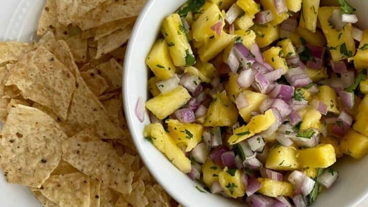 A white bowl of pineapple salsa with red onion and cilantro sits on a plate next to a pile of tortilla chips.