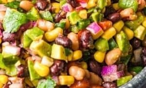 A close-up of a colorful salad with black beans, corn, avocado, red onion, tomatoes, cilantro, and green bell pepper in a speckled bowl.