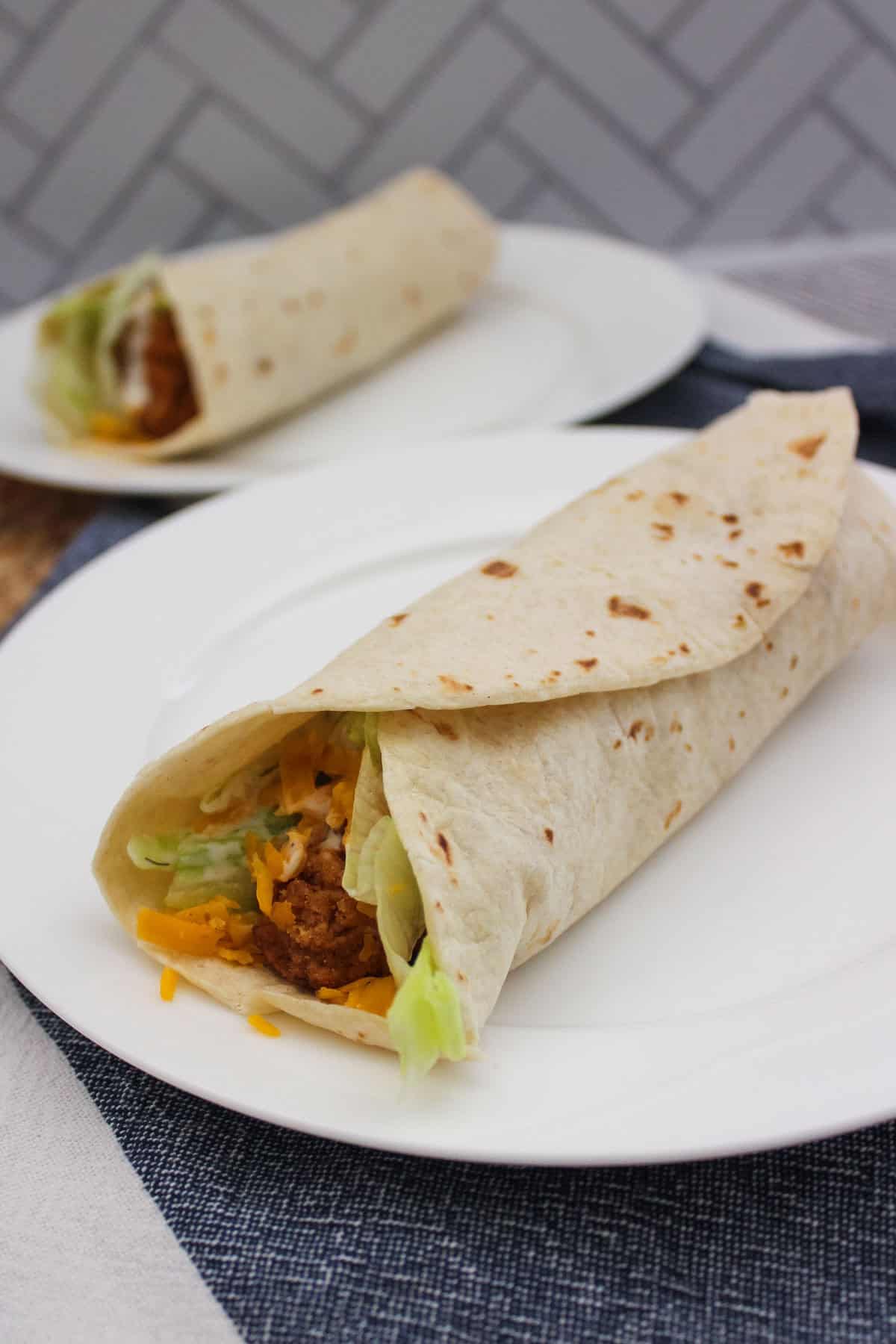 A close-up of two soft tortilla wraps filled with breaded chicken, shredded lettuce, and cheddar cheese, served on white plates with a patterned background.
