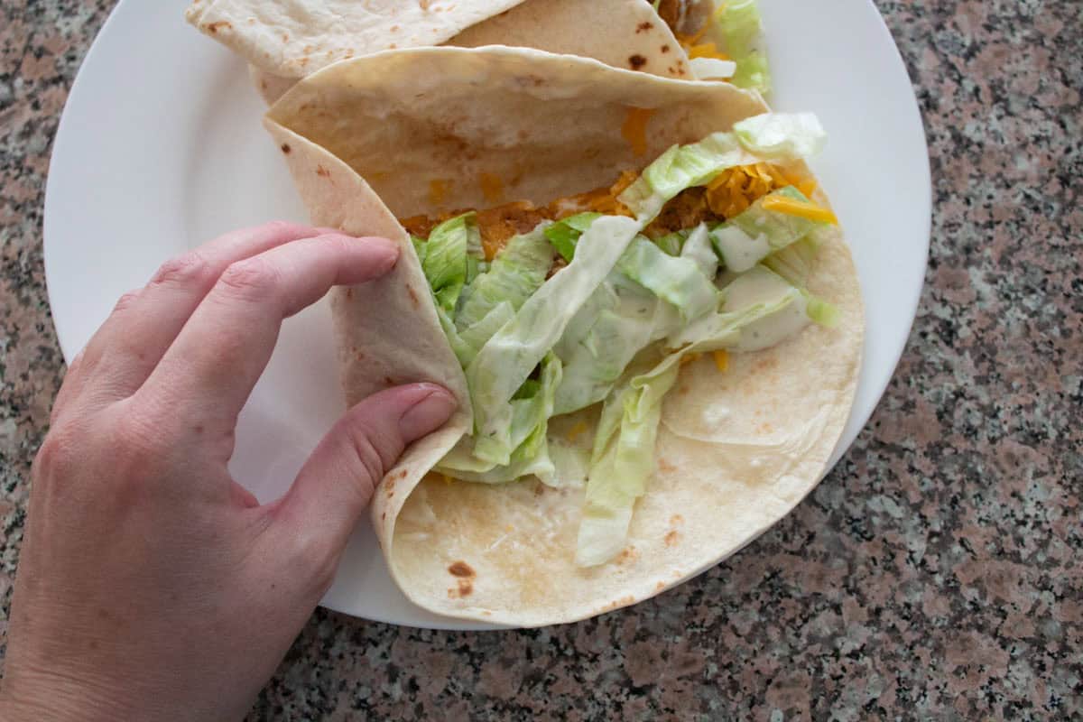 A hand holding a tortilla with lettuce, cheese, chicken, and sauce on a flour tortilla. Another wrap sits on a white plate, all on a granite countertop.